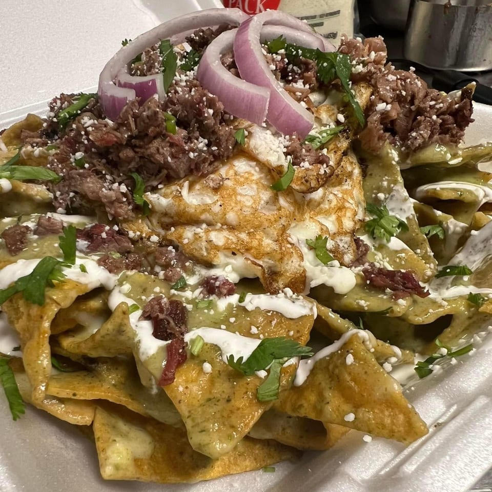 Chilaquiles with shredded beef, fried egg, red onion, cilantro, and crema in a white container.