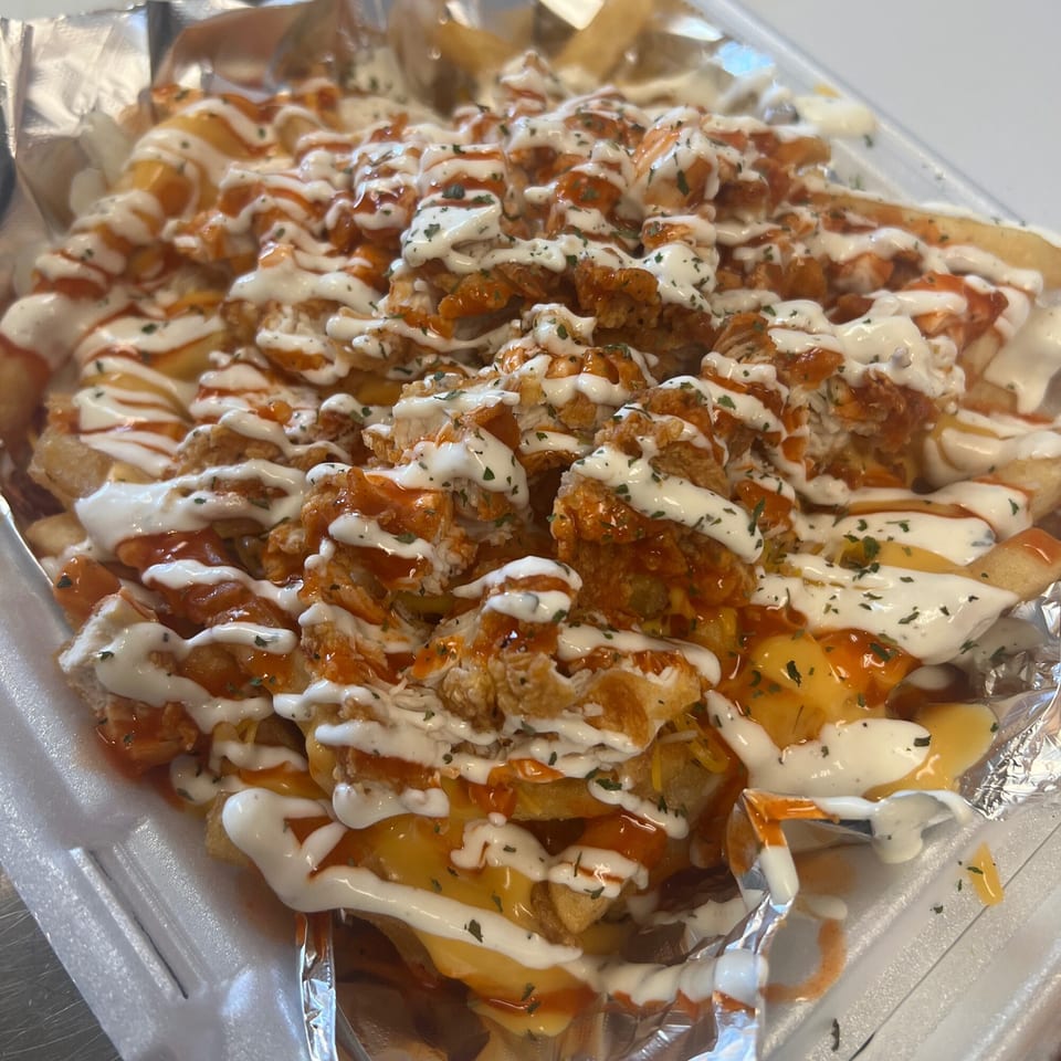 Loaded fries with crispy chicken, melted cheese, white sauce, and hot sauce in a foil-lined container.