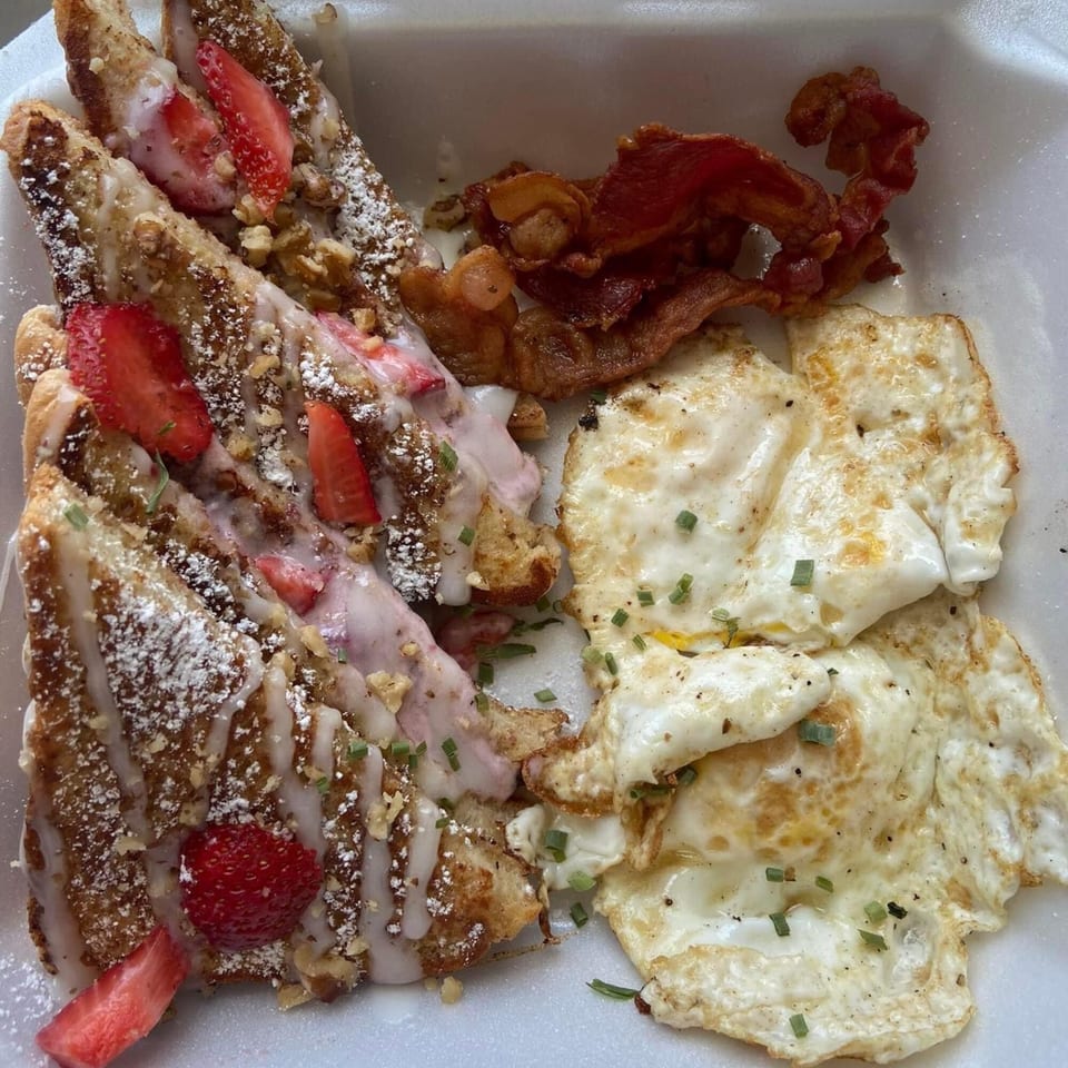 French toast with strawberries, bacon, and fried eggs with chives in a white takeout container.