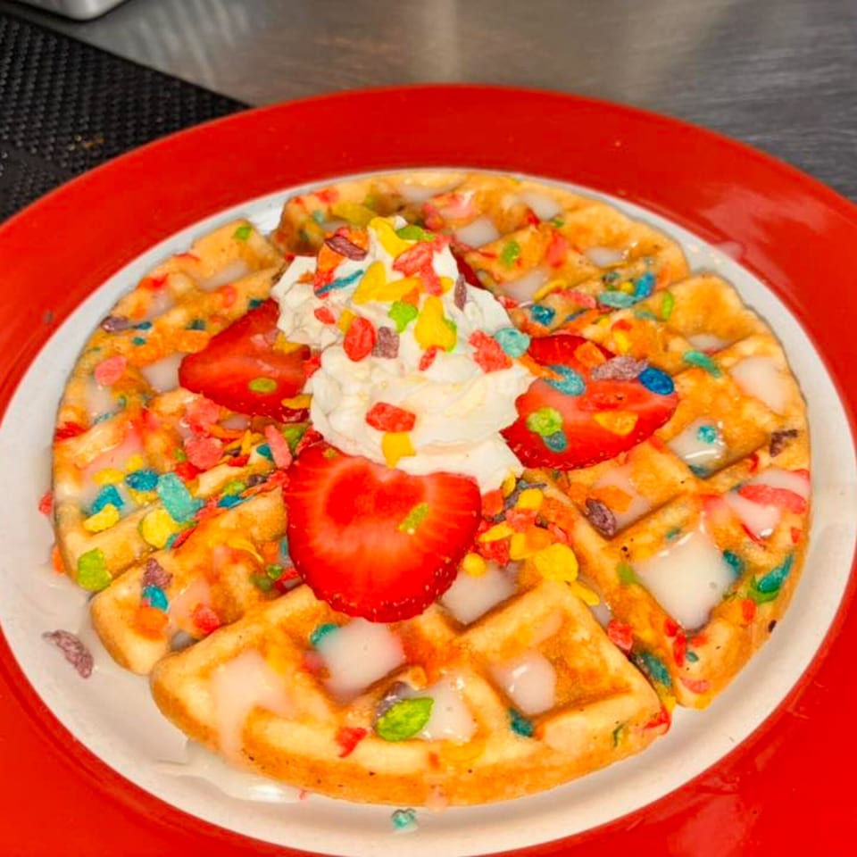 A waffle topped with whipped cream, strawberries, colorful cereal, and white icing on a red plate.