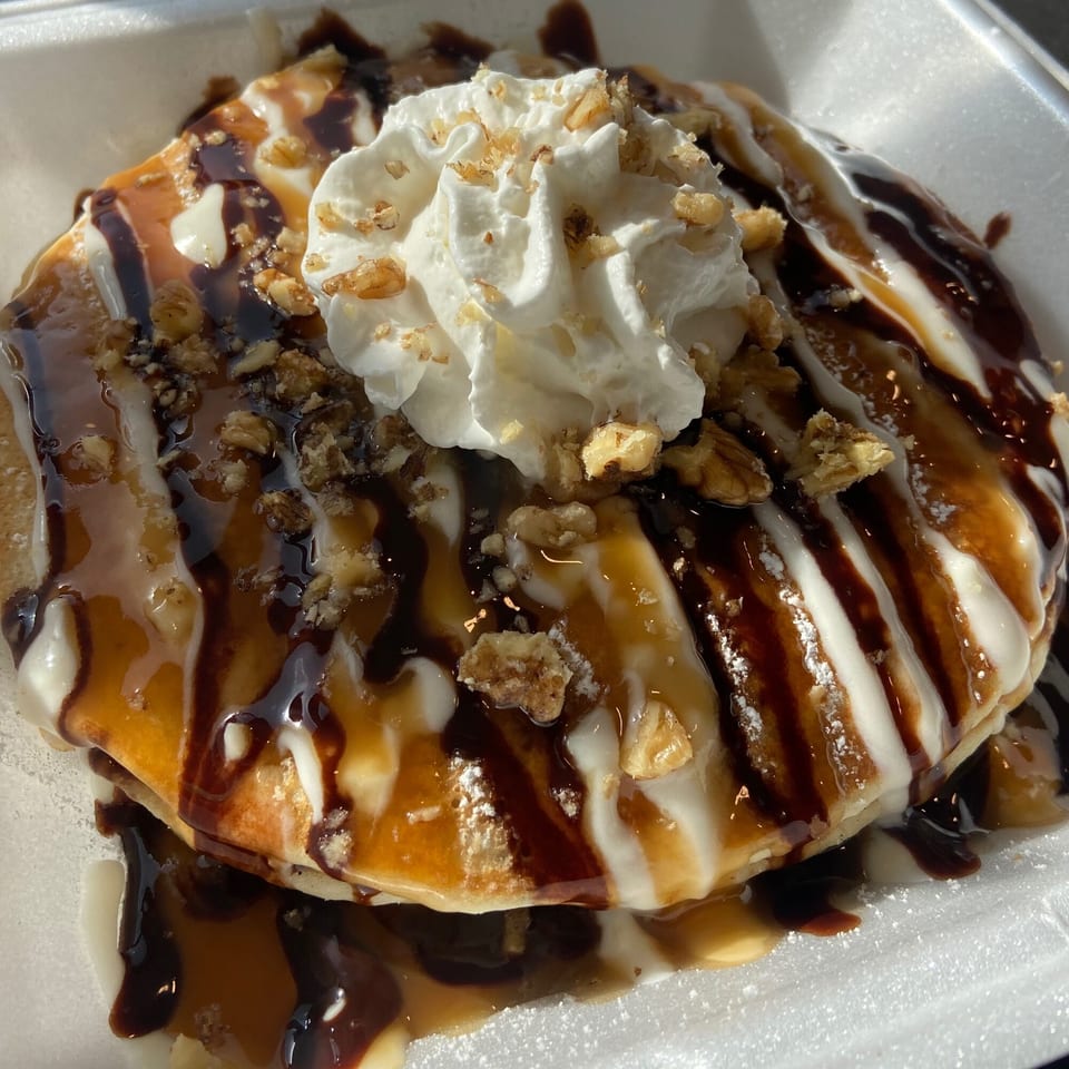 Pancakes topped with whipped cream, chocolate, caramel, and nuts in a white takeout container.