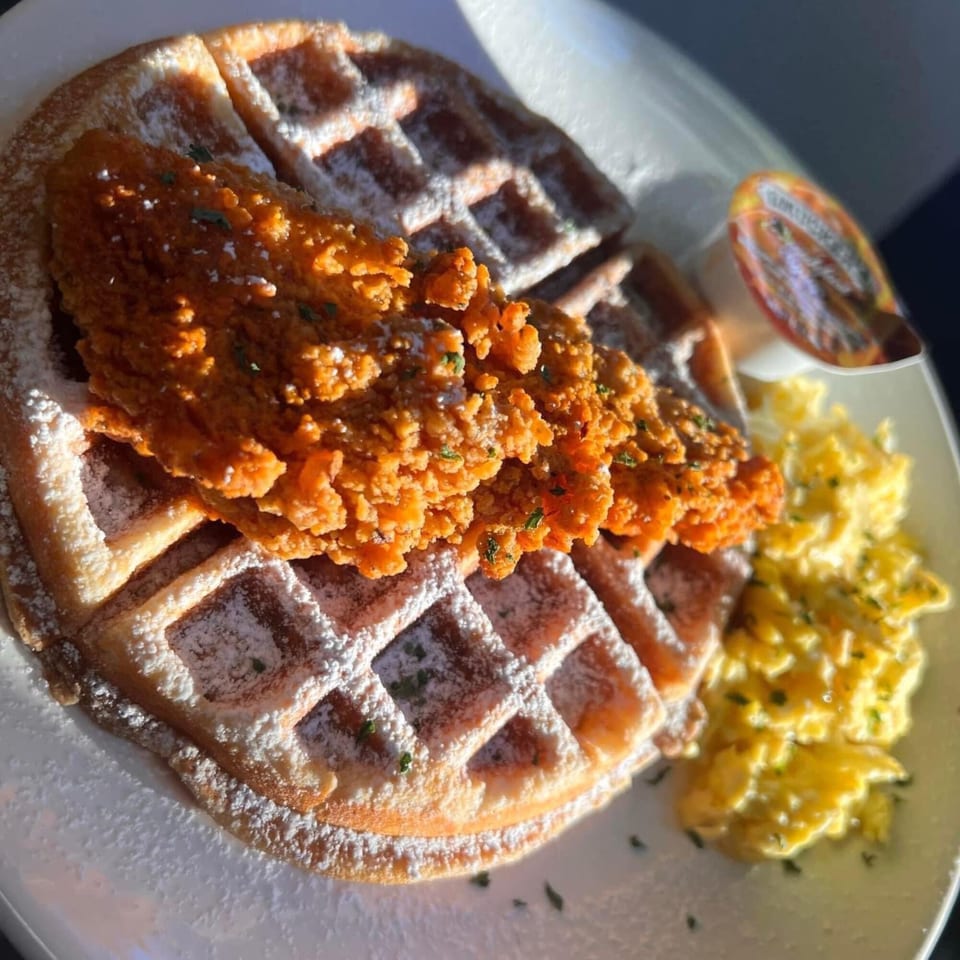 Crispy fried chicken on a powdered sugar waffle with scrambled eggs and syrup on a white plate.